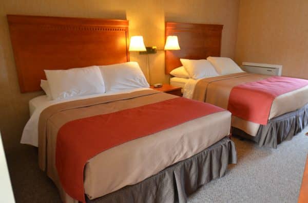 The image shows a hotel room with two beds featuring white linens and red blankets, wooden headboards, bedside lamps, and a warm color scheme.