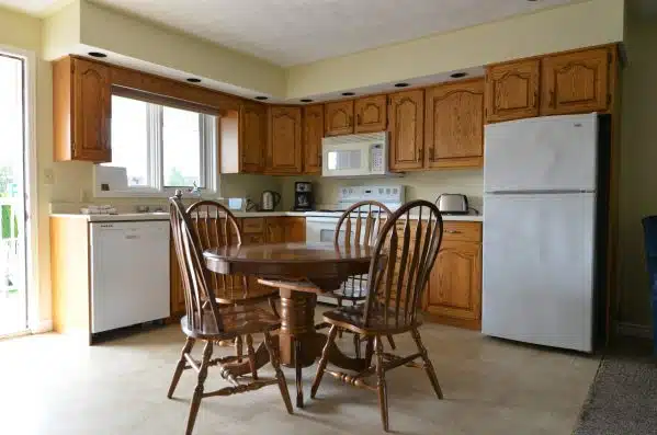 A spacious kitchen with wooden cabinetry, white appliances, and a round dining table with matching chairs. Natural light comes in through a window by the sink.