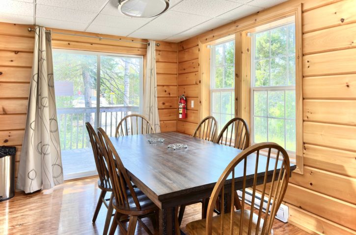 Wooden dining room with table and six chairs. Large windows and sliding door offer natural light. Fire extinguisher mounted on wall.