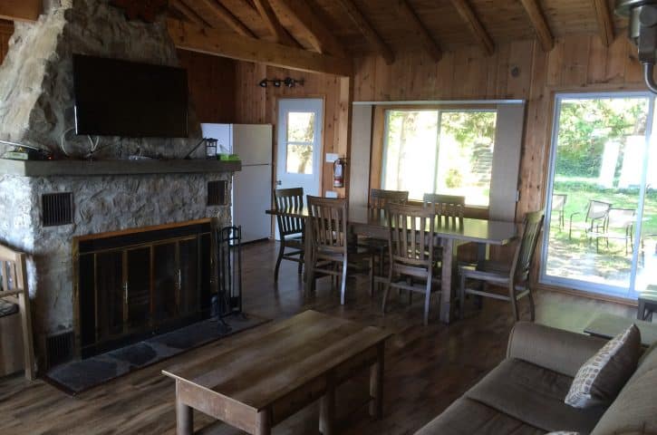 A cozy rustic living room with a stone fireplace, wooden furniture, couch, and dining area, surrounded by large windows with an outdoor view.