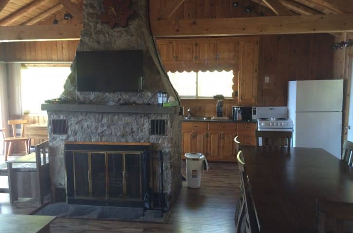 Rustic cabin interior with a stone fireplace, wooden walls and ceiling beams, a TV above the mantel, and a kitchen with wooden cabinets.