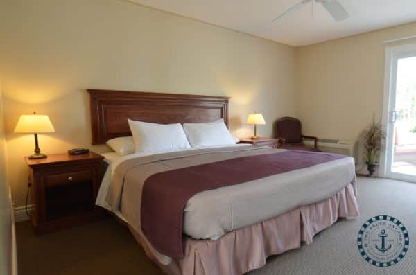 The image shows a neatly arranged bedroom with a large bed, two bedside tables, lamps, a fan overhead, and a door leading outside.