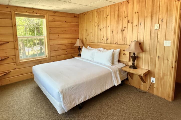 A cozy wooden bedroom features a neatly made bed with white linens, two lamps, and a window offering a view of greenery.