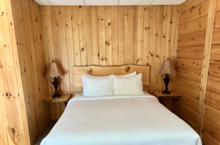 Cozy wooden cabin bedroom with a neatly made bed, white sheets, elegant bedside lamps, and contrasting textures in the walls and carpet.