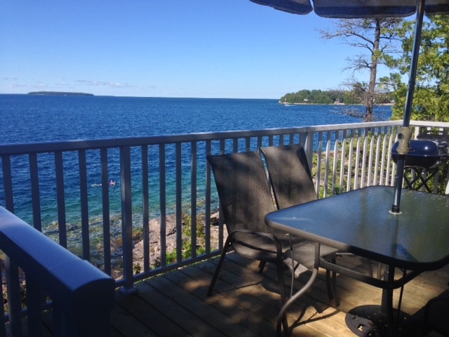 A serene waterfront view from a deck with a railing, outdoor furniture, and a clear sky, overlooking a calm blue lake with distant islands.