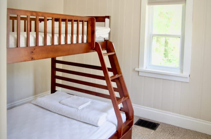 A small, bright room features a wooden bunk bed with white bedding, next to a window with a view of greenery.