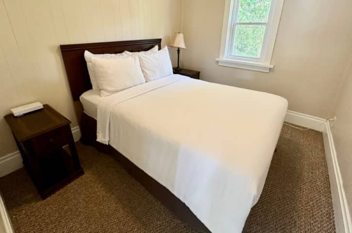 A cozy bedroom with a neatly made bed, white linens, nightstand, lamp, and window with a view of greenery.