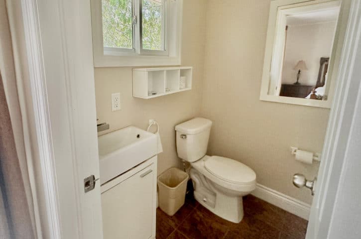 A small bathroom features a sink, toilet, mirror, and window. The room has beige walls, a wooden floor, and minimalistic design.