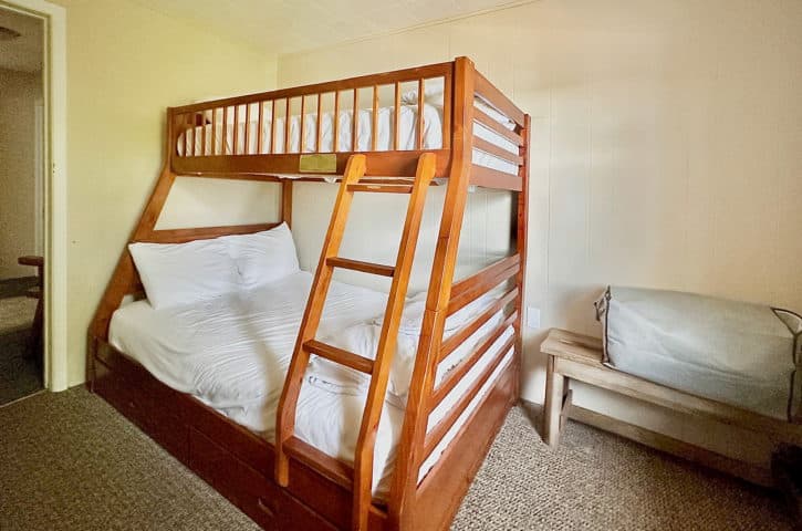 A cozy bedroom features wooden bunk beds with white bedding, a small wooden bench, and beige walls, creating a simple, inviting space.