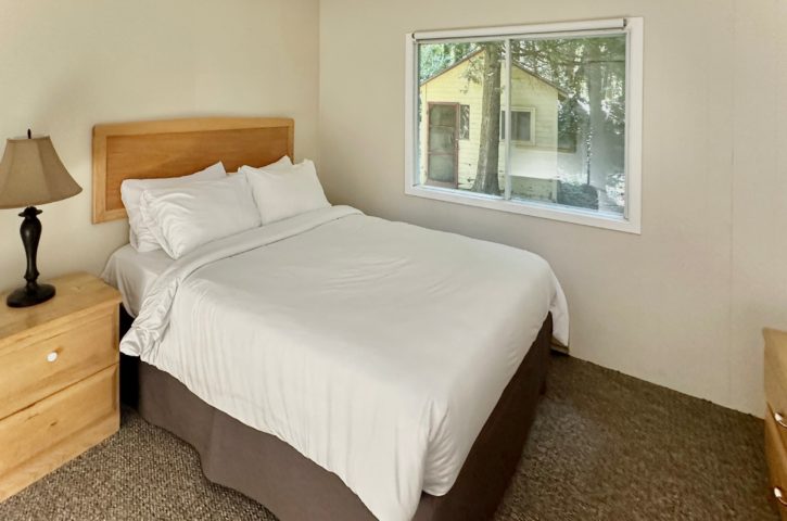 A cozy bedroom with white bedding, wooden furniture, and a lamp. A window reveals a small cabin and trees outside.