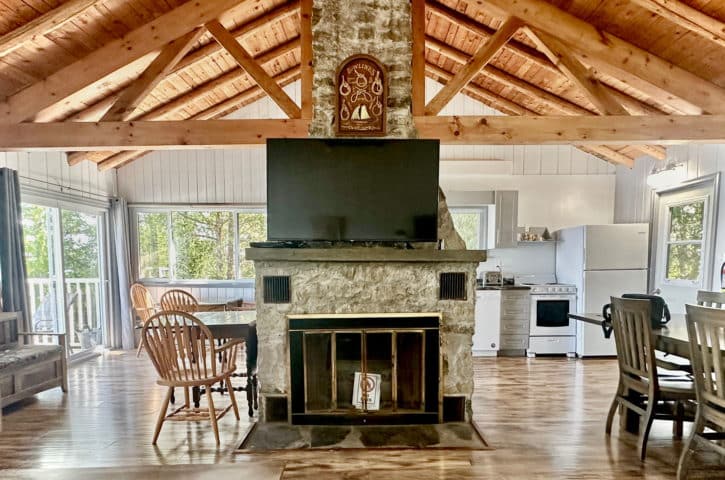 A cozy, rustic cabin interior with wooden beams, stone fireplace, modern kitchen, and dining area. Large windows show lush greenery outside.