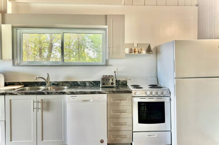 A cozy kitchen with white cabinets, a marble countertop, stove, dishwasher, and refrigerator. Sunlight filters through a window, highlighting woodland views.